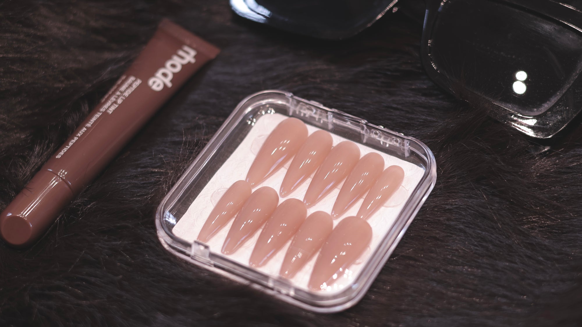 Nude coffin press-on nails in a clear case on dark fur with lip tint and sunglasses